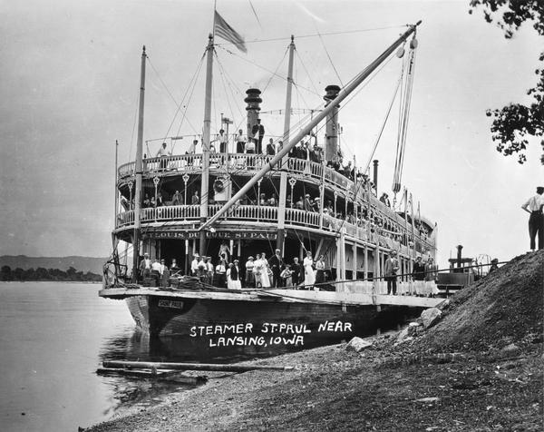 The bow of sidewheel excursion, "Saint Paul," at shore. Passengers are on all three decks. Sign on bow reads: "St. Louis, Dubuque, St. Paul." Later named the "Senator."