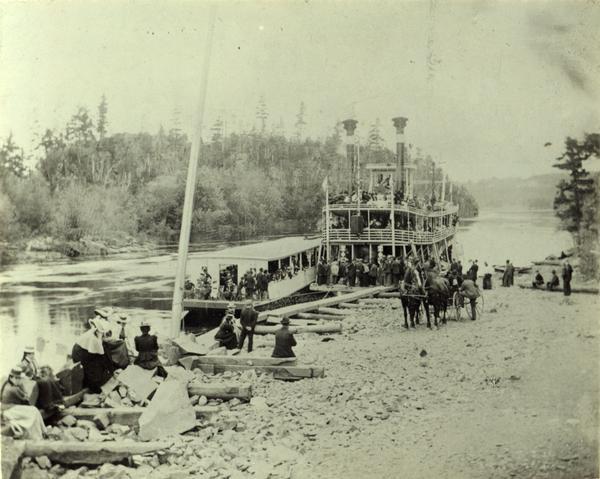 A fire fighter's excursion on the "Vernie Mac" at a landing near Taylor Falls on the St. Croix River between Minnesota and Wisconsin.  Excursion barge is along side and people are seated watching from the bank. A team of horses is in the foreground.