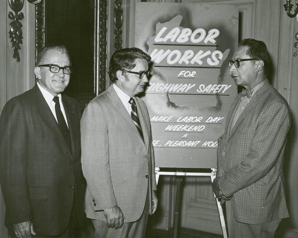 Three men, including Governor Patrick J. Lucey, pose in front of a sign that reads: "Labor Works For Highway Safety. Make Labor Day Weekend A Safe, Pleasant Holiday". The sign was part of a public relations campaign that called attention to the increased need for safety on holidays when many vacationers were on the highways.