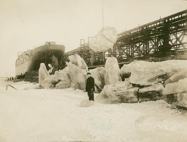 A man is standing in front of huge blocks of ice at the American Shipbuilding Company. The ship "War Otter" is in the background. A large chunk of ice tied with rope is suspended in the air just behind the man.