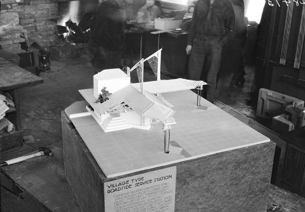 Architectural model of a village type roadside service station designed by Frank Lloyd Wright. Men are standing behind the model, and there is a fire in the fireplace behind the men.