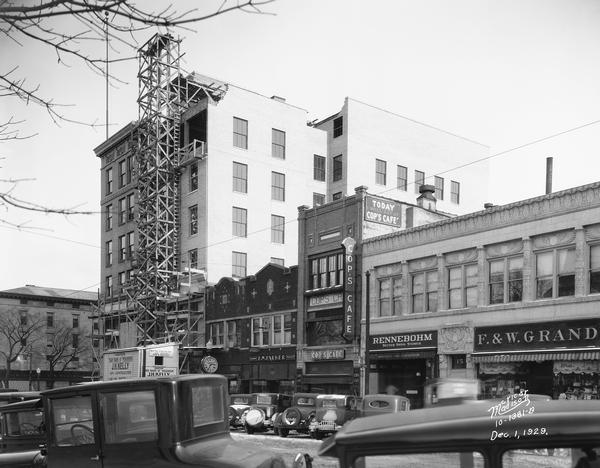 Scaffolding rises on the State Bank of Wisconsin, 1 West Main Street, as an addition is constructed. Automobiles are parked on both sides of the street in front of Woolworths (1 E. Main aka Pioneer Building), Badger Candy Kitchen (7 W. Main), E.W. Parker Jewelers (9 W. Main), Cop's Cafe (11 W. Main), Rennebohm's drugstore (13 W. Main), and the Levitan Building (15 W. Main).