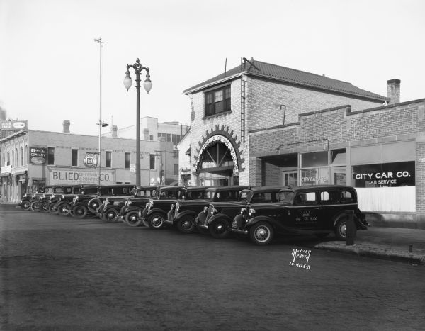 A fleet of cars is lined up in front of City Car Company Cab Service located at 214 East Washington Avenue. Building on the left became Paradise Printing, 206 East Washington Avenue. Building on the right became Pahl Tire in 1936. Pahl Tire moved to 202 E. Washington Avenue ca. 1960 and closed in 2013.
