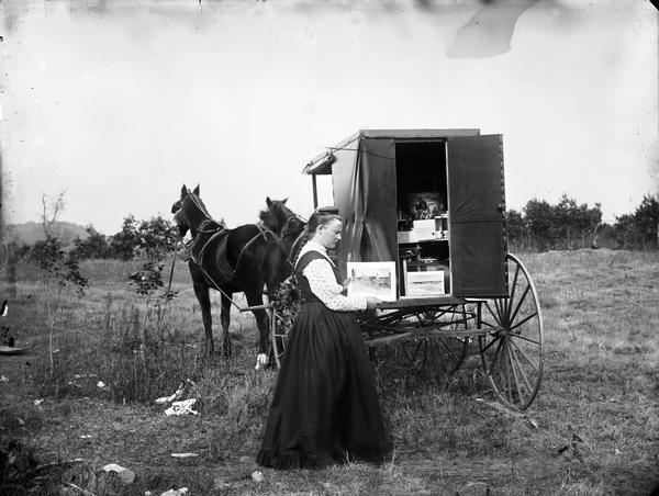 A woman, possibly the daughter of C.R. Monroe, holding up an image of Black River Falls Main Street at the back of a traveling photographer's wagon.