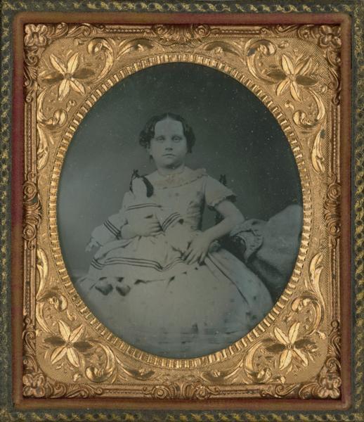 Sixth plate ferrotype/tintype portrait of Anna Martin Gourlay (1858-1864). She is seated, facing forward, wearing a short-sleeved dress, and holding a doll in her right arm. Hand coloring on dress neckline and sleeves. Gold details applied to necklace.<p>Annie was born April 7, 1858 in Glens Falls (Warren Co.), New York, the daughter of Andrew Gourlay (1785-1866), a tailor, and Jane Jack (1815-1899). Annie died in Glens Falls on November 24, 1864. She was buried at Glens Falls Cemetery.<p>Photo taken by "Conkey" on January 5, 1864. Conkey was probably George W. Conkey (1837-1910), who was born in New York City, and in 1861 came to Glens Falls, where he opened a photography studio and gallery.</p>