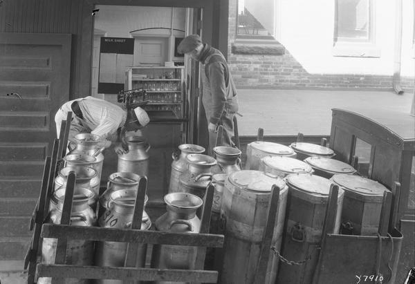 Two men deliver milk in cans to a creamery from the back of a milk truck.