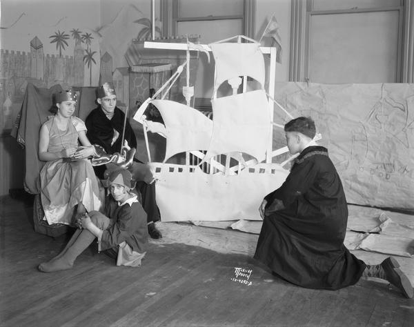 Four children in costume surround a cardboard cutout of a sailing ship for a play about Christopher Columbus. The play was performed at Longfellow School, 1002 Chandler Street in the Greenbush neighborhood. The actors from left to right include Rose Bonnino, Angelo Caruso, Ernest La Bella, and Paul Cuccia.