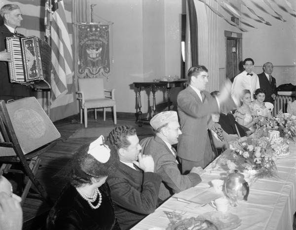 "Jobo" Joe Puccio, leads the singing at the Italian Women's Mutual Society 7th anniversary banquet in the Italian Workingman's Club, 914 Regent Street, in the Greenbush neighborhood. In the crowd are George Salerno playing the accordion; Tony Parrish, standing in the white shirt with a bow tie, Theodore Paratore, seated on Tony's left, and Arthur Olsen, American Legion commander.