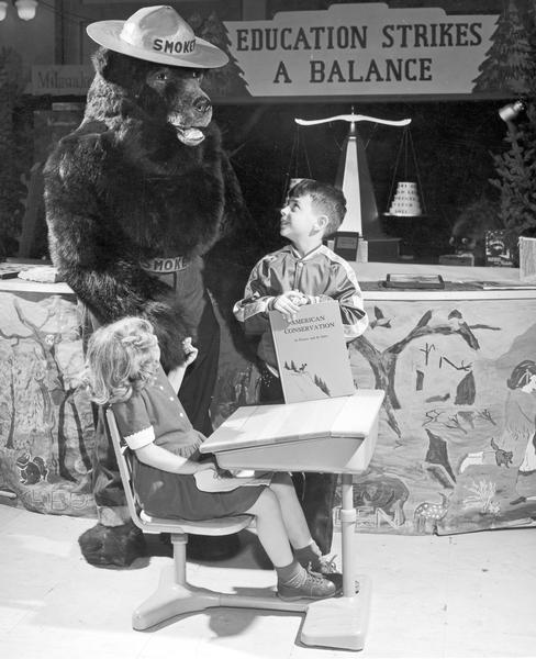 A man dressed in a Smokey Bear costume is standing with two Milwaukee school children. A young girl is sitting at a desk, and a boy is standing and holding a book titled: "American Conservation." Both children are looking up at Smokey Bear. Behind them in what may be an exhibit is a sign above a set of scales that reads: "Education Strikes A Balance."