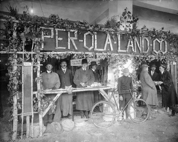 Men in hats and coats stand in exhibit booth for the Per-Ola Land Company.