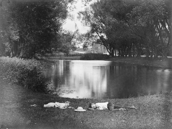 Two boys lying on the grass asleep in Tenney Park near the lagoon with a hat on the ground between them. The Sherman Avenue bridge and the Hausmann Brewery malt house are in the background.