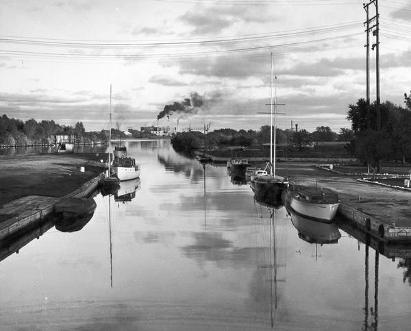 Harbor in Marinette at sunset. Boats are docked at the sides of a canal. Smoke pours from smokestacks of a facility in the distance.