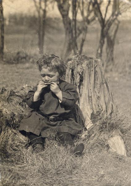 James Peterson, son of the photographer, playing the harmonica. He is sitting against a stump, with trees in the distance.