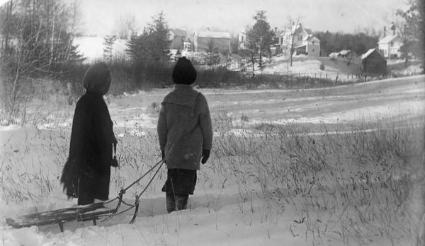 Winter scene with Muriel and Laurie Peterson, the photographer's children, reflecting on the snow conditions near their home. Laurie holds the rope of a sled.