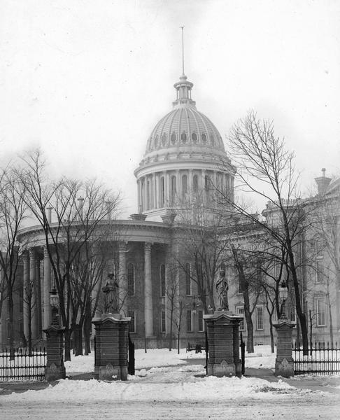 Third Wisconsin State Capitol (the second in Madison), showing the East Washington Avenue gateway and iron fence. In reality the statues, which were meant to represent the four seasons, were less impressive than they seem in vintage photographs. They were made of cast zinc that was painted white, not stone sculptures.