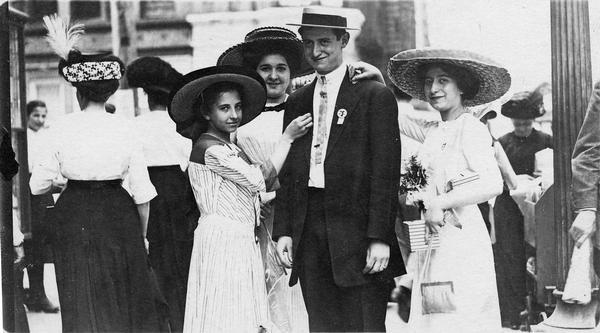 A man in a hat and jacket poses with three well-dressed women wearing dresses and wide-brimmed hats.