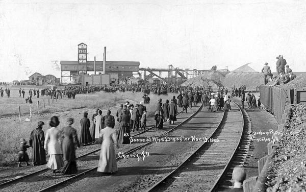 Photographic postcard of an elevated view of people walking to the mine (in the distance) after a disaster. Caption reads: "Scene at Mine Disaster, Cherry, Ill."