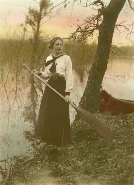 Woman standing on riverbank with canoe paddle and boat moored at tree.