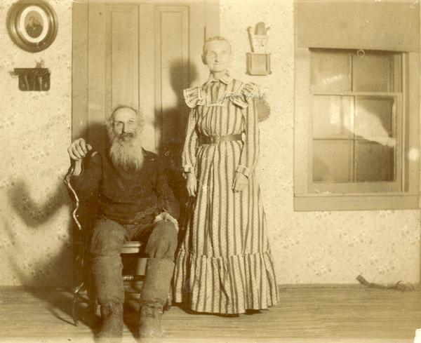 Julius Porter Wait and his wife, Lucretia Melvina (Mosher) Wait. He has a beard, and is sitting on a chair, wearing dark work clothes and tall, loose boots, and holding a crooked wooden walking stick or cane. She is standing, wearing a long-sleeved, floor-length dress of striped cotton, with a small round collar and shoulder ruffles; the dress is belted at the waist with a dark belt with a metal buckle.