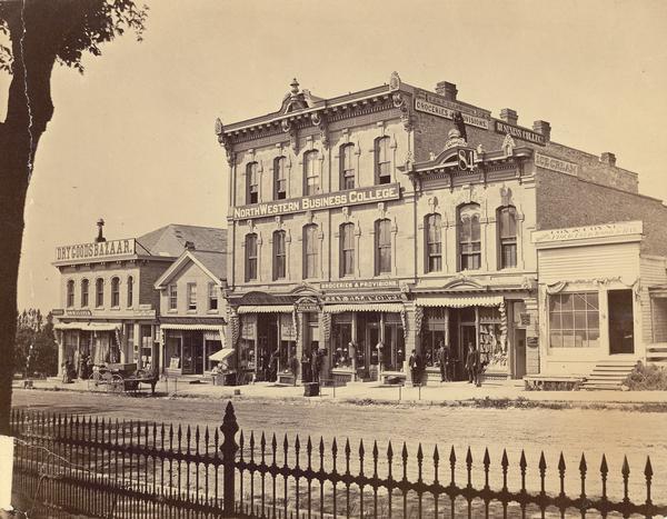 Ellsworth Block on North Pinckney Street between East Washington and East Mifflin Streets as seen from the Capitol Park. Storefronts include the Dry Goods Bazaar, Singer Sewing Machines, the Hollister & Wittman Pharmacy, the Ellsworth Grocery Store, and the Northwestern Business College. Men are standing in front of the buildings near a horse-drawn wagon. The foreground provides a close-up of the iron fence constructed around the Capitol Park that was erected in 1873. As constructed, the fence represented a less expensive version of the original designed by Stephen V. Shipman, the architect who also designed the Capitol dome.