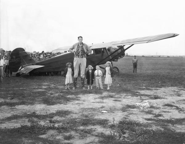 A giant and other circus performers, including Harry and Daisy Earles, posing in front of a Midwest Air Transport airplane while a crowd is watching from behind the airplane.