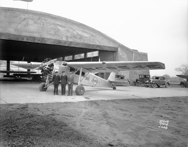 Two airplane pilots and a third man are standing in front of a Northwest Airways air mail plane at Madison's Royal Airport. There are automobiles parked near a hangar in the background.