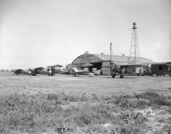 Midwest Air Transport planes in front of the hangar at the Madison Airport.  Located northeast of the city near the Oscar Mayer plant, the field was sometimes referred to as North Street Airport.  Pilots of the Midwest Air Transport Service are posed in front of each airplane.  Also in clear view is the tower of the airport's 24-inch rotating beacon.  The Midwest Air Transport Service was out of business by 1933. 
