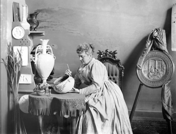 Studio portrait of Susan Frackelton, sitting at a table painting a bowl and surrounded by her art work and a large medal. Caption on back reads: "Mrs. Susan S. Frackelton of Milwaukee, WI. Artist in ceramics and inventor of the gas kiln, frequent exhibitor of decorative pottery and medal winner at leading expositions in the United States and Europe in the 1880s and 1890s. <i>Chicago Times Herald</i>, Sunday, Feb. 17, 1901."