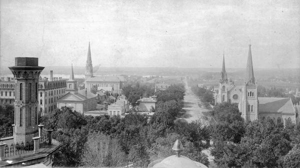 Elevated view from the Wisconsin State Capitol dome toward West Washington Avenue. The Park Hotel is on the left. The four church steeples that can be identified are, left to right, First Baptist Church, St. Raphael Catholic Church, First Congregational Church, and Grace Episcopal Church. The left foreground provides a close view of the decoration on a Capitol chimney.