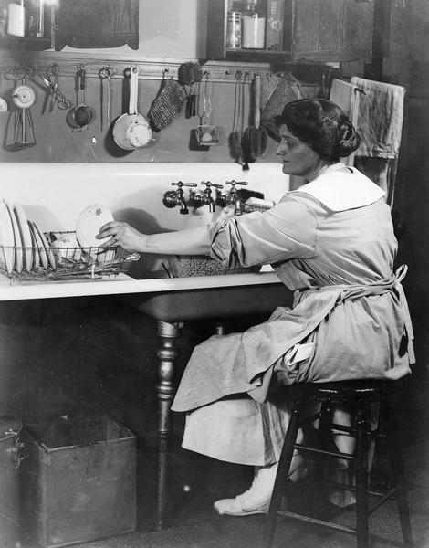 View of a woman wearing an apron sitting on a stool and washing dishes at a kitchen sink. Kitchen utensils are hanging on the wall above her.