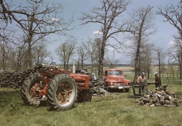 Father and son at work cutting wood with McCormick Farmall H tractor running belt-driven circular saw on rural farm. Behind them is parked an International truck.