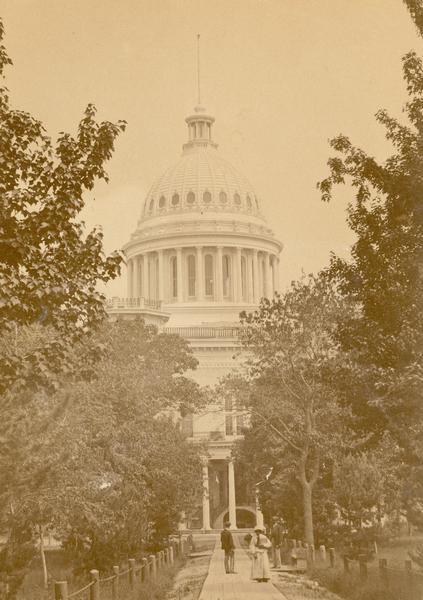 The third Wisconsin State Capitol as it appeared during the early 1870s. Close scrutiny of the entrance (whether it is the north or south entrance cannot be determined), reveals a semi-circular staircase under the portico that is nowhere else so visible in the historical documentation.  In 1873 the rustic wooden walkways and chain fence gave way to a more designed landscape.