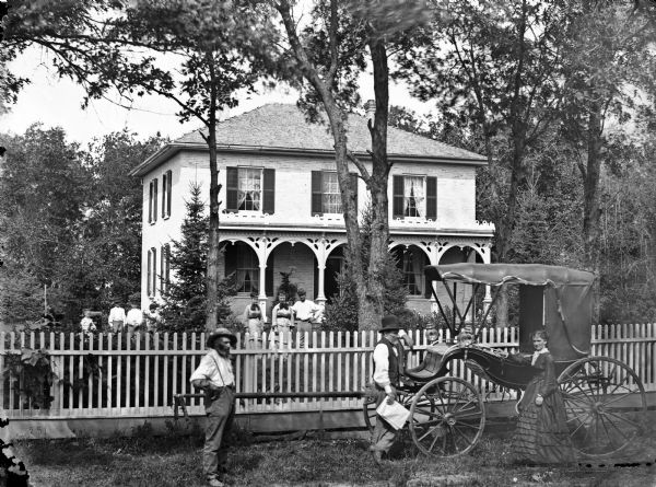 An elegant carriage and a group of people are standing in front of a picket fence, with a brick house with shutters and hipped roof behind. More people are standing in the yard behind the fence.