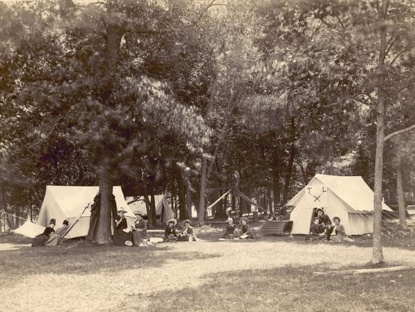 A well-dressed group of campers picnic outside their tents (perhaps late 1800's).