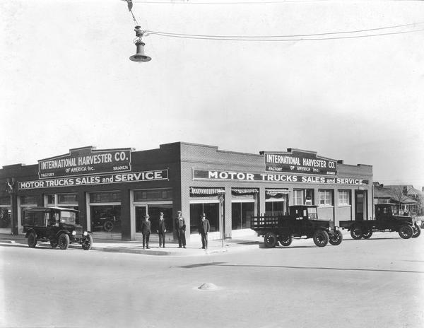 International Harvester motor truck dealership. Four men are standing on the sidewalk in front of the building. Three International trucks are parked in the street.