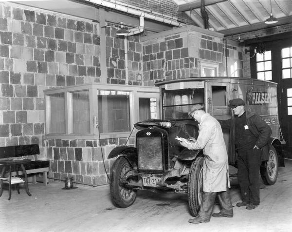 Mechanic with clipboard and trouble light examines the engine of an International truck used to deliver "Holsum" brand bread at an International Harvester motor truck branch service station. The driver of the truck is looking on.