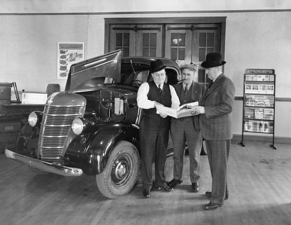 Showroom of International Harvester general line and motor truck dealership of Herr Brothers in Elrose(?), Saskatoon, Canada. Three men are standing together looking at an International truck advertising brochure next to a new International truck. The men are (left to right) Jack Herr, W.H. Boerjan (farmer) and G.L. Elliott, branch manager at Saskatoon. A literature rack is against the back wall.