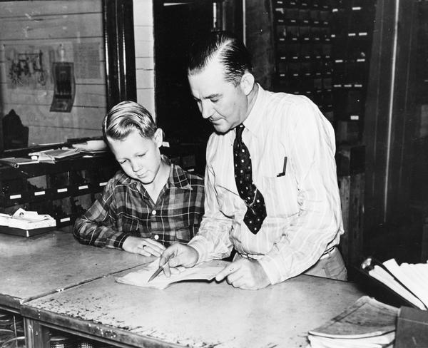 International Harvester dealer pointing out something in an open booklet to a young boy. Original caption appears to read: "dealer security."