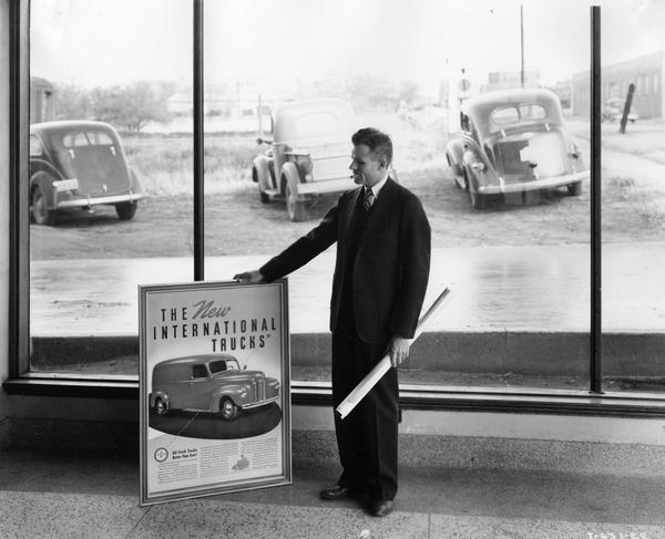 Man with a cigar in his mouth standing in front of a showroom window holding a framed poster for "the new International trucks." Two cars and a truck are in the parking lot outdoors.