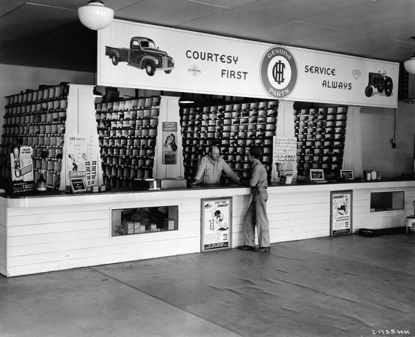 Two men talking at the sales counter of an International Harvester dealership. Advertisements adorn the counter and parts bins are in the background. The dealership sold both trucks and farm equipment.