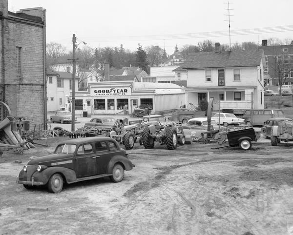 Farm machinery on dealer's side lot,  with car in the foreground and Good Year tire store across the street.