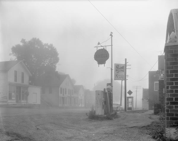Street scene with a Shell gasoline station, the Kitty Korner Kafe, and a phone booth in early morning fog.