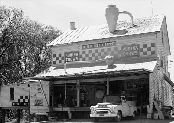 A front view of the Mielke Feed & Supply grist mill, with Purina Chows signs placed above the loading dock area.  A GMC pickup truck is  parked in the loading area.