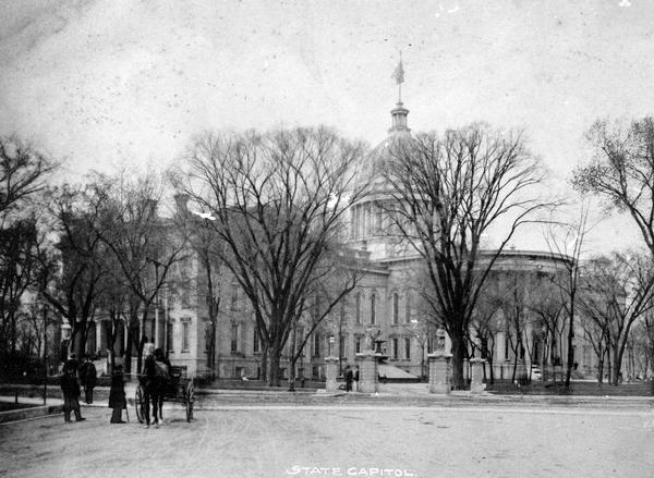 Pedestrians and a horse and cart at the  intersection of Main Street and Monona Avenue (South Wisconsin Avenue until December 1, 1877; Martin Luther King Jr. Boulevard as of January 19, 1987). Behind the stone entrance gate, the centennial fountain under its winter covering can be seen.