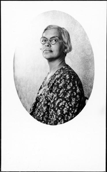 Waist-up studio portrait of Lillie Rosa Minoka-Hill. She is wearing round spectacles.