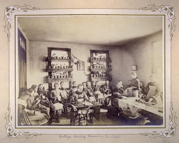 "Cottage" sewing room at the Wisconsin Industrial School for girls, depicting a class of young girls handling fabrics, sewing, and working with sewing machines.