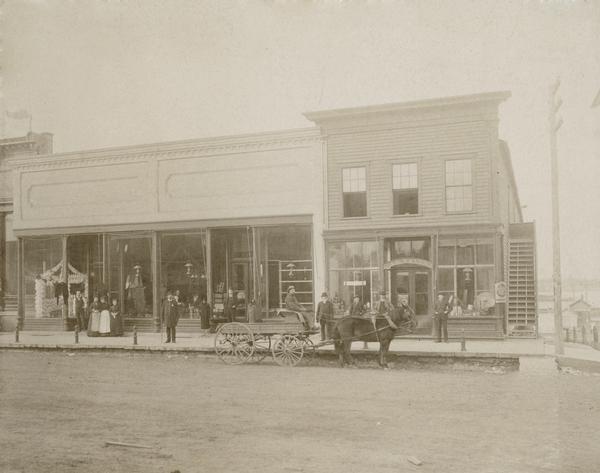 People pose on sidewalk in front of businesses including A.J. Wells. There is a horse-drawn wagon parked at the side of the road.