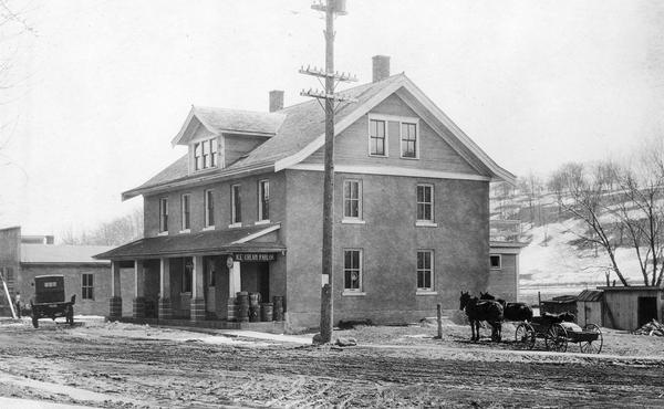 Exterior of ice cream parlor with a horse-drawn wagon parked on the right with sheds behind. Barrels are stacked on the porch. Signs above the porch read: "Ice Cream Parlor" and "Coca-Cola". On the left is a truck and another building. The street is dirt with a sidewalk along the edge. In the background is a hill partially covered with snow.