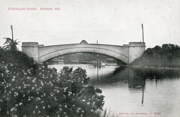 View from shoreline towards the Steensland Bridge with a boat and passengers passing beneath. Caption reads: "Steensland Bridge, Madison, Wis."