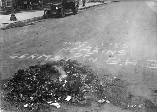 "Here lies the remains of German in B.H.S." is written on the pavement next to a smoldering pile of German textbooks. A car is parked along the curb in the background.
